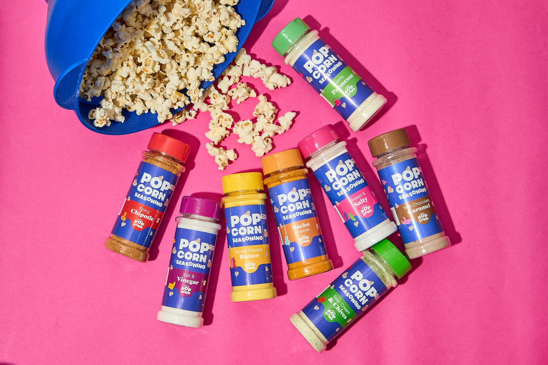 Popcorn spilling from a blue Microwave Popper Bowl beside nine Pass The Popcorn seasoning bottles on a pink background