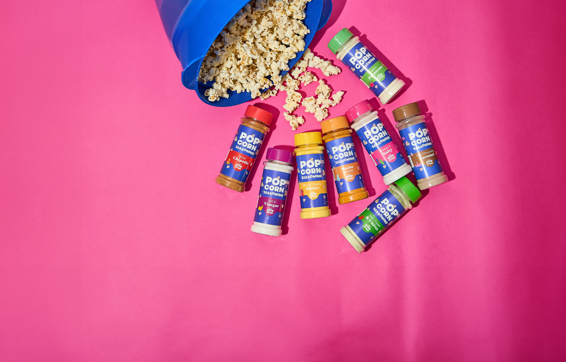 Pass The Popcorn seasoning bottles laid out with popcorn and kernels spilling across a pink background